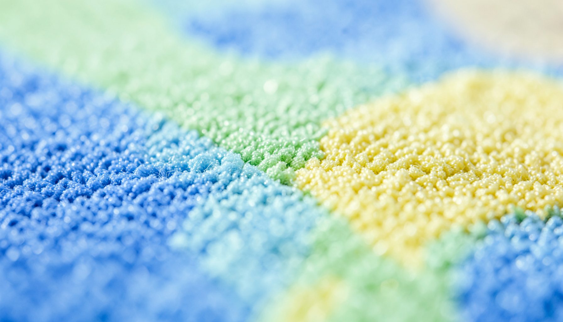 Gros plan extrême sur la texture d'un tapis de jeu coloré en mousse, aux tons pastel de bleu, vert et jaune, avec une surface douce et légèrement granuleuse captée en lumière naturelle.