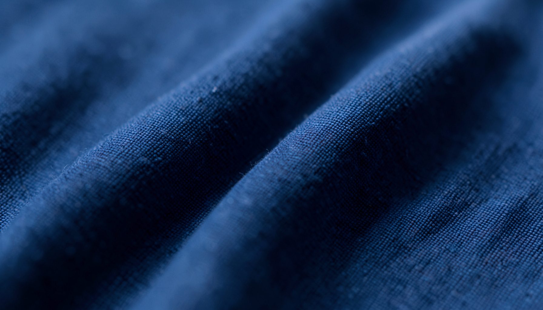 Gros plan extrême sur un tissu en coton bleu marine doux, légèrement froissé, avec des reflets chauds et une texture mate et accueillante.